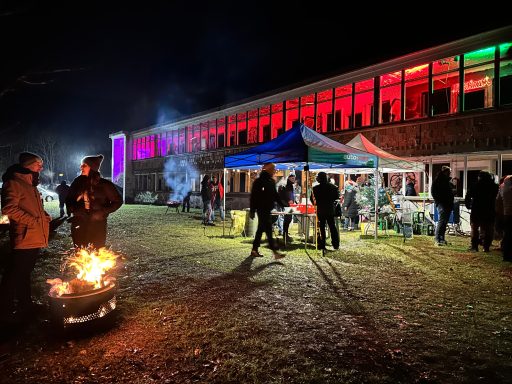 Menschen versammeln sich bei Nacht an einem Gebäude mit bunten Lichtern und Feuerstelle.